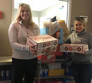 Two women holding boxes of donuts in front of pantry items for donation